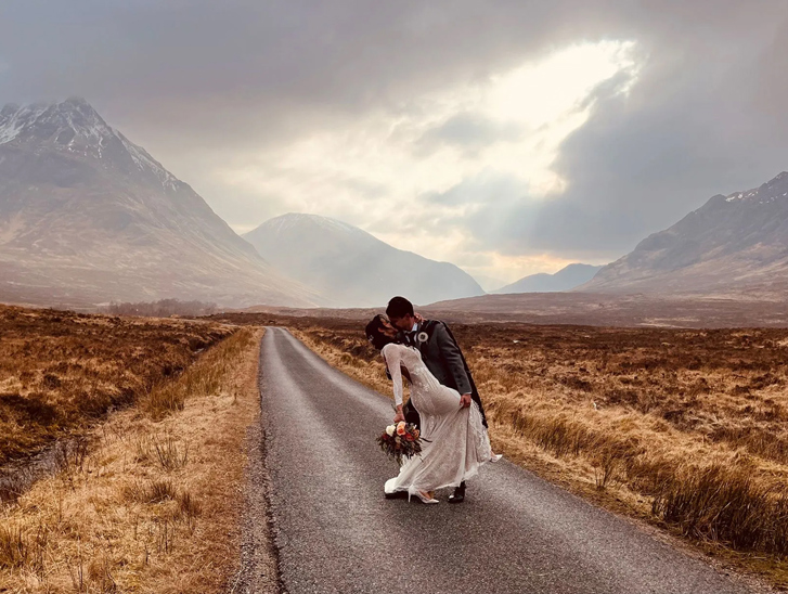 Groom dips bride holding bouquet in kiss with snow covered mountain tops in background